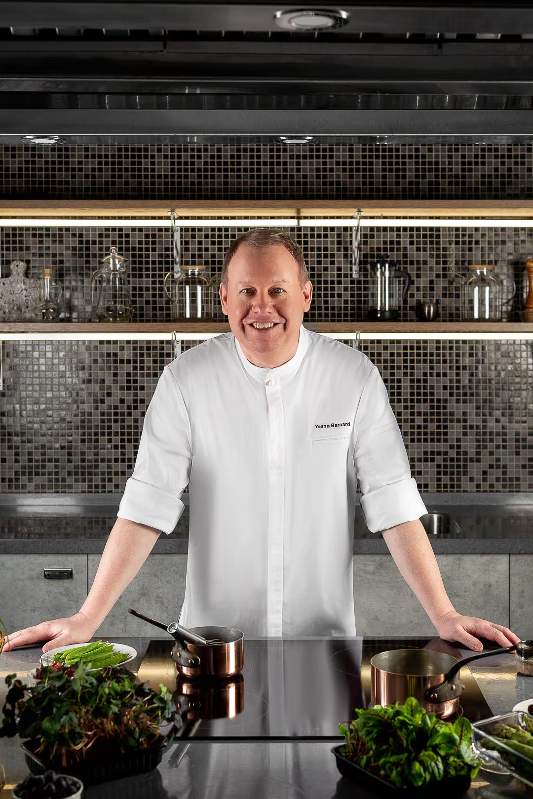 Chef Yoann Bernard souriant en veste de cuisine dans sa cuisine professionnelle, entouré d'herbes fraîches et de casseroles en cuivre